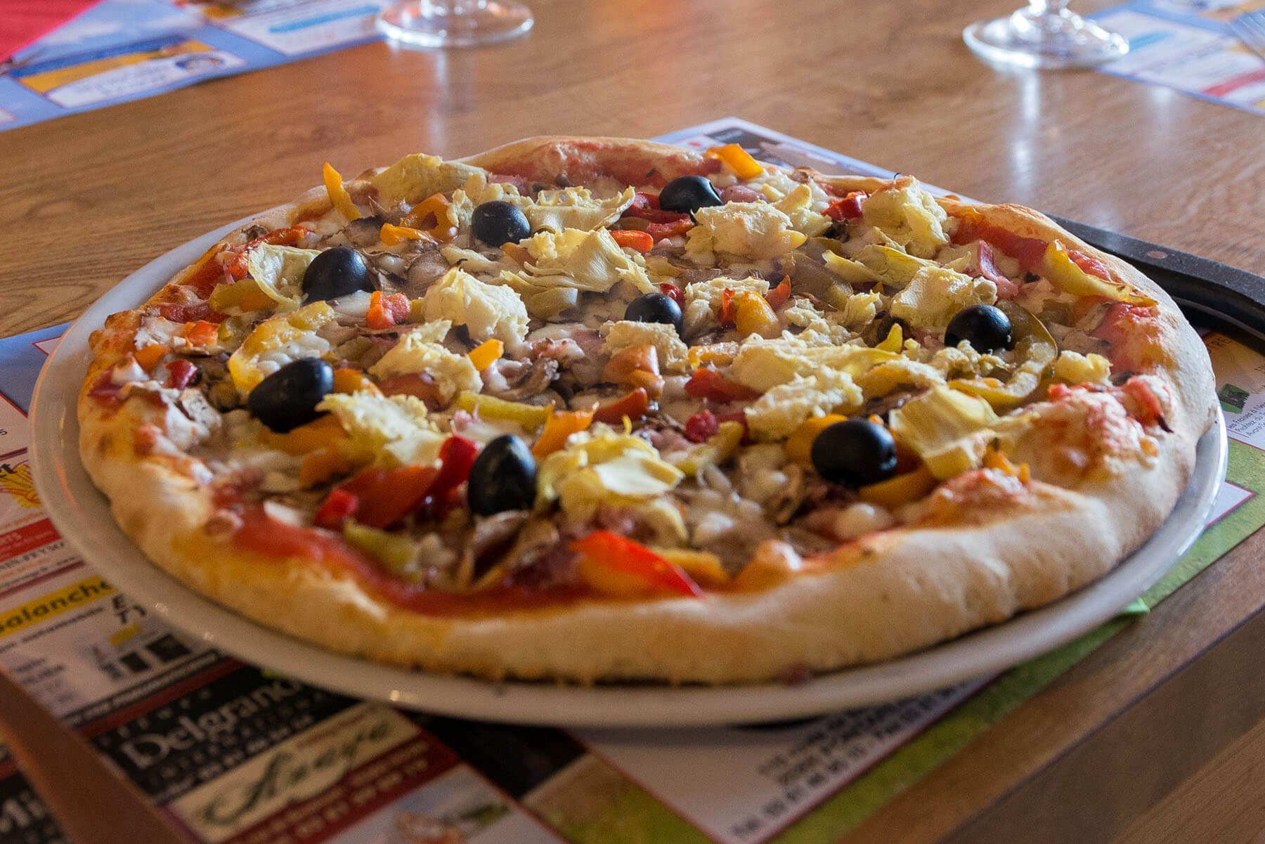 Pizza sur une assiette blanche, garnie d'olives, d'artichauts et de légumes, sur une table de restaurant.