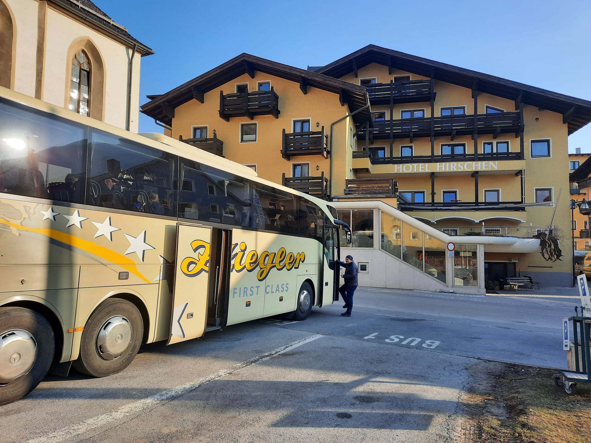 Bus parked in front of a yellow hotel with balconies. A person stands near the bus. Sunny, outdoor setting.