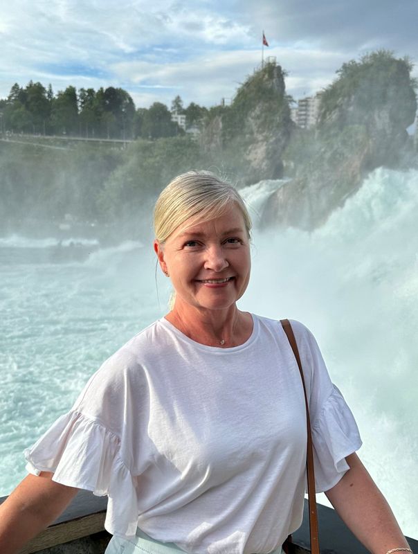 Lächelnde Frau vor dem Rheinfall in der Schweiz. Sie trägt ein weißes Oberteil, im Hintergrund der Wasserfall.