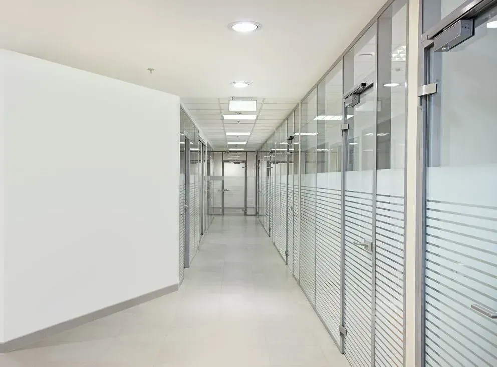 Langer, heller Büroflur mit Räumen mit Glaswänden und mattierten unteren Paneelen, weißen Wänden und einer abgehängten Decke.