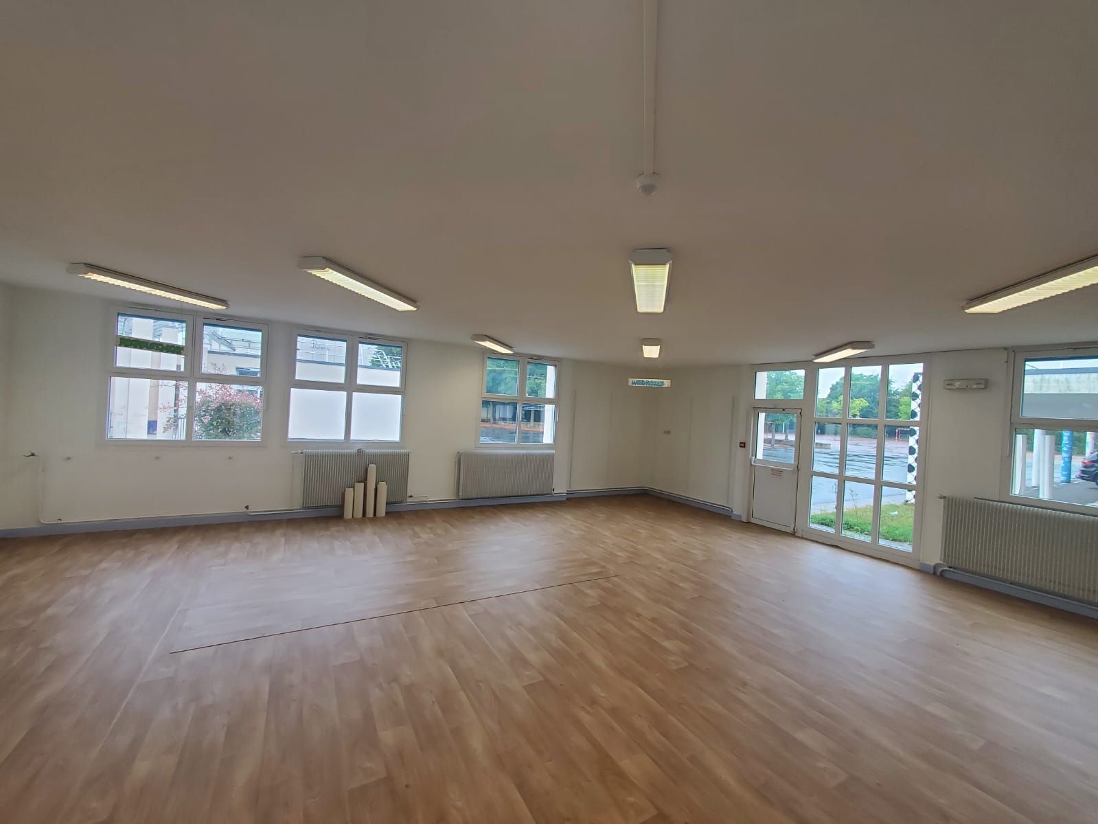 Pièce lumineuse et vide avec parquet, murs blancs, fenêtres et éclairage fluorescent au plafond.