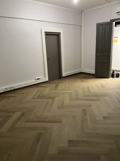 Pièce vide avec parquet à chevrons, murs blancs et deux portes sombres.
