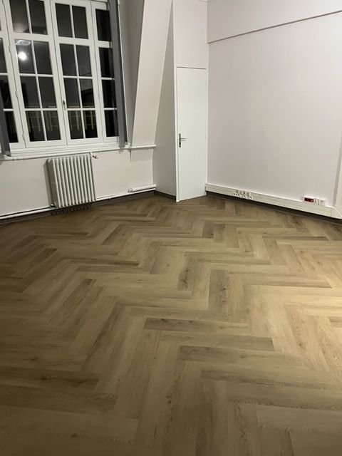 Pièce vide avec parquet à chevrons, murs blancs, fenêtre, radiateur et porte ouverte