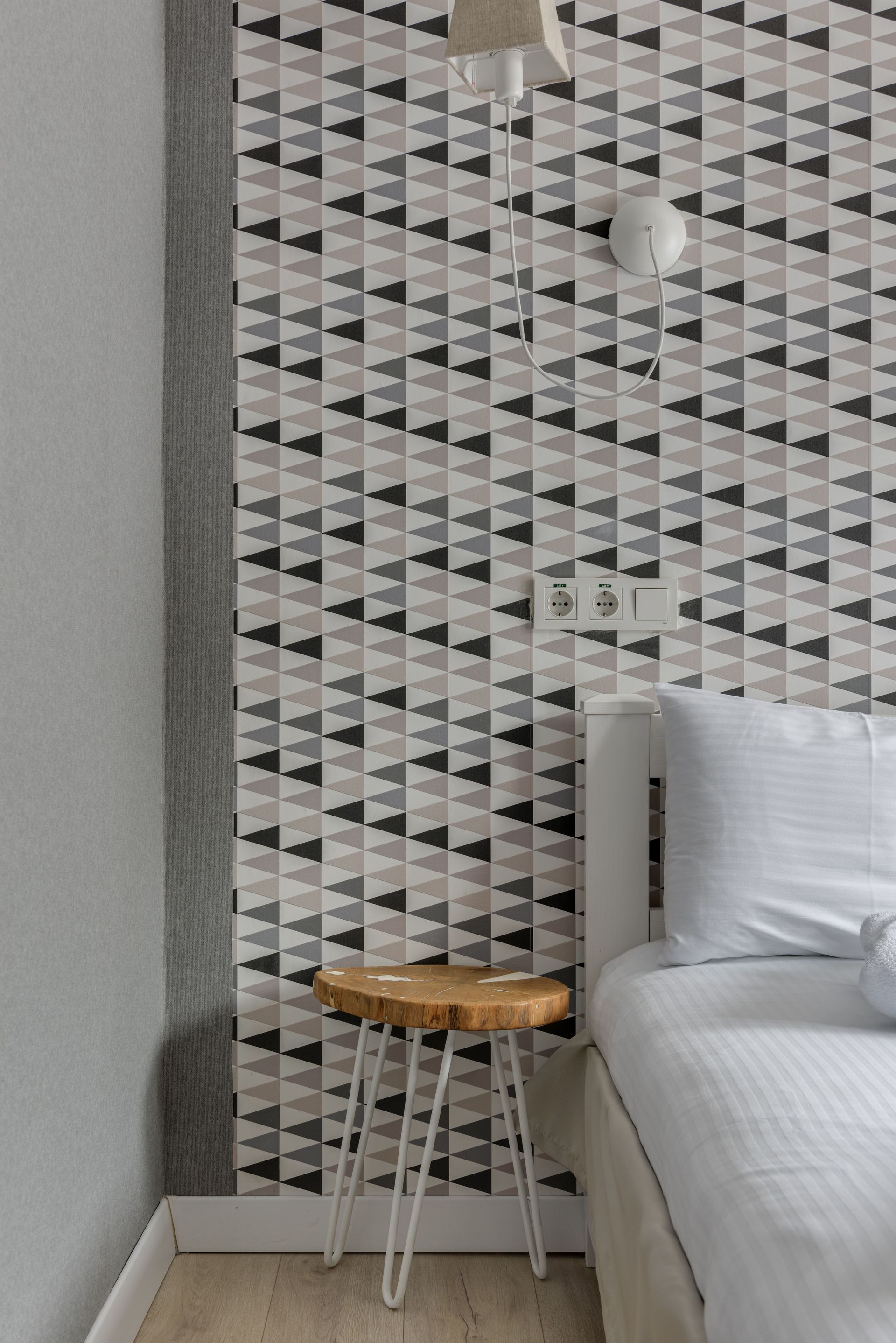 Coin de chambre moderne avec papier peint à motifs, lit blanc et petit tabouret en bois à pieds en métal.