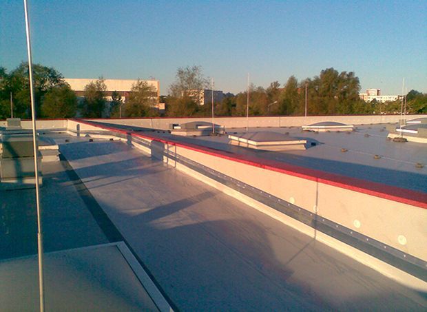 Dachterrasse mit rotem Sicherheitsgeländer, Oberlichtern und Blick auf ferne Gebäude unter blauem Himmel.