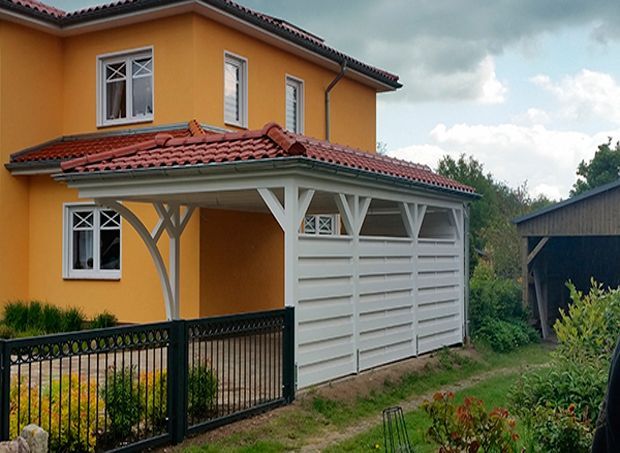 Gelbes Haus mit weißem Carport; schwarzer Zaun, rote Dachziegel und grüner Rasen.