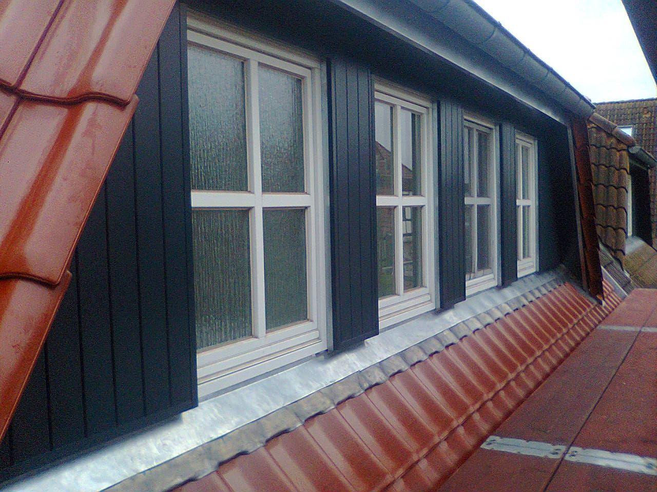 Eine Reihe weißgerahmter Fenster an einer dunkel getäfelten Wand vor einem roten Ziegeldach.