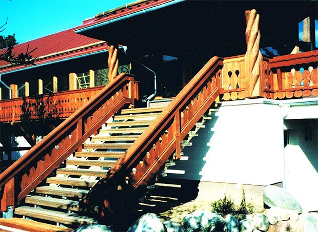 Eine hölzerne Außentreppe führt hinauf zu einem Gebäude mit rotem Dach, überdachter Veranda und braunem Geländer.