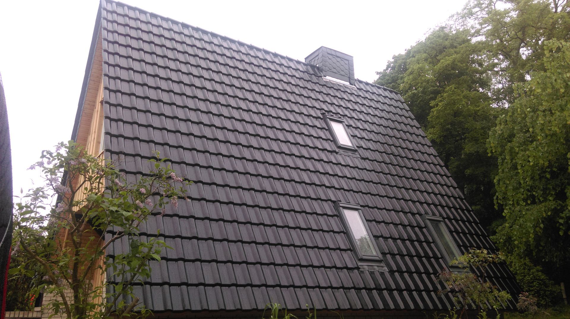 Schwarzes Ziegeldach eines Hauses mit zwei Dachfenstern, einem Schornstein und Bäumen in einer bewölkten Tageslandschaft.