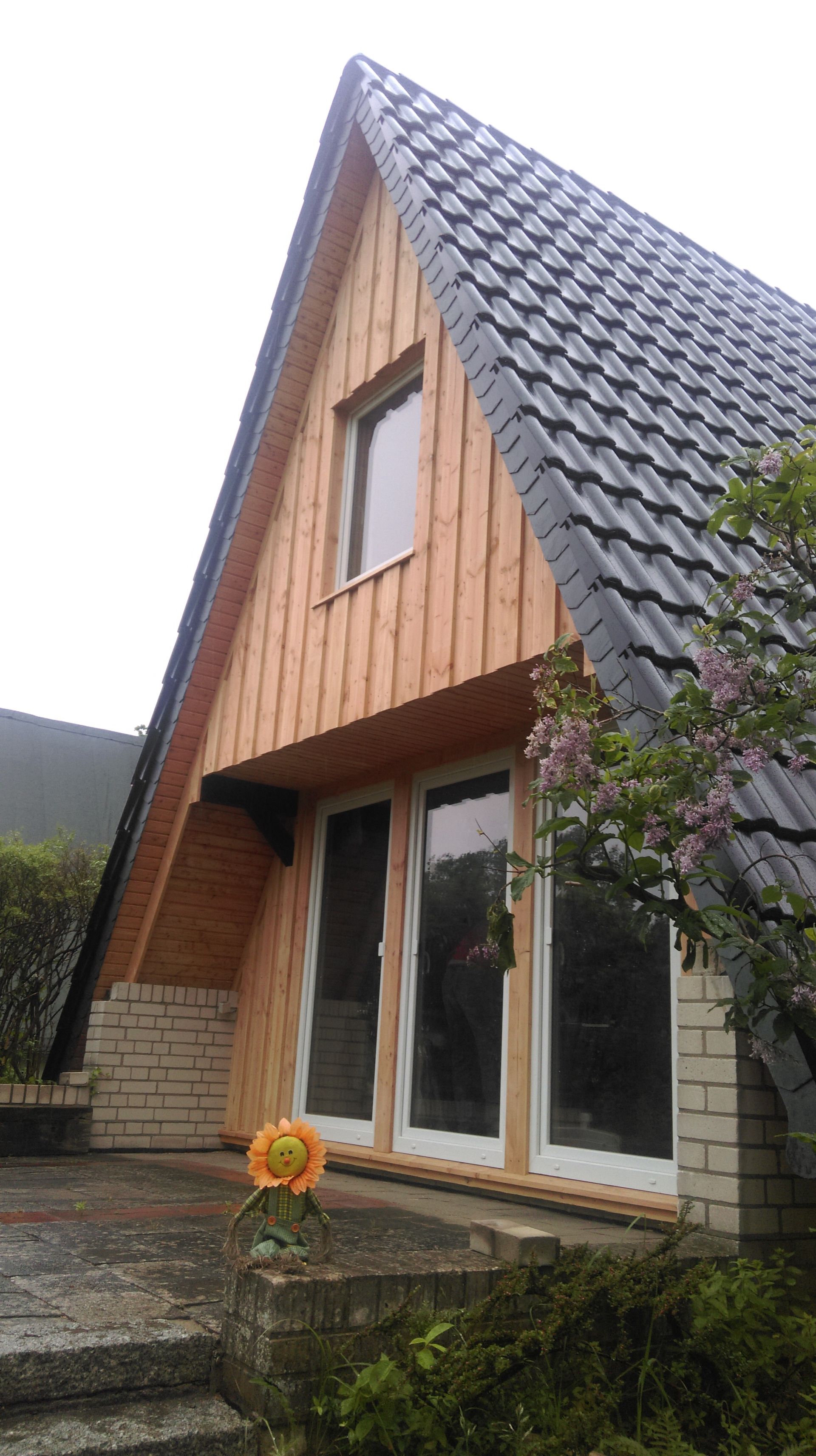 Ein A-förmiges Haus mit Holzfassade, dunklem Dach, Ziegelsockel und Glastüren; davor blüht eine Blume.