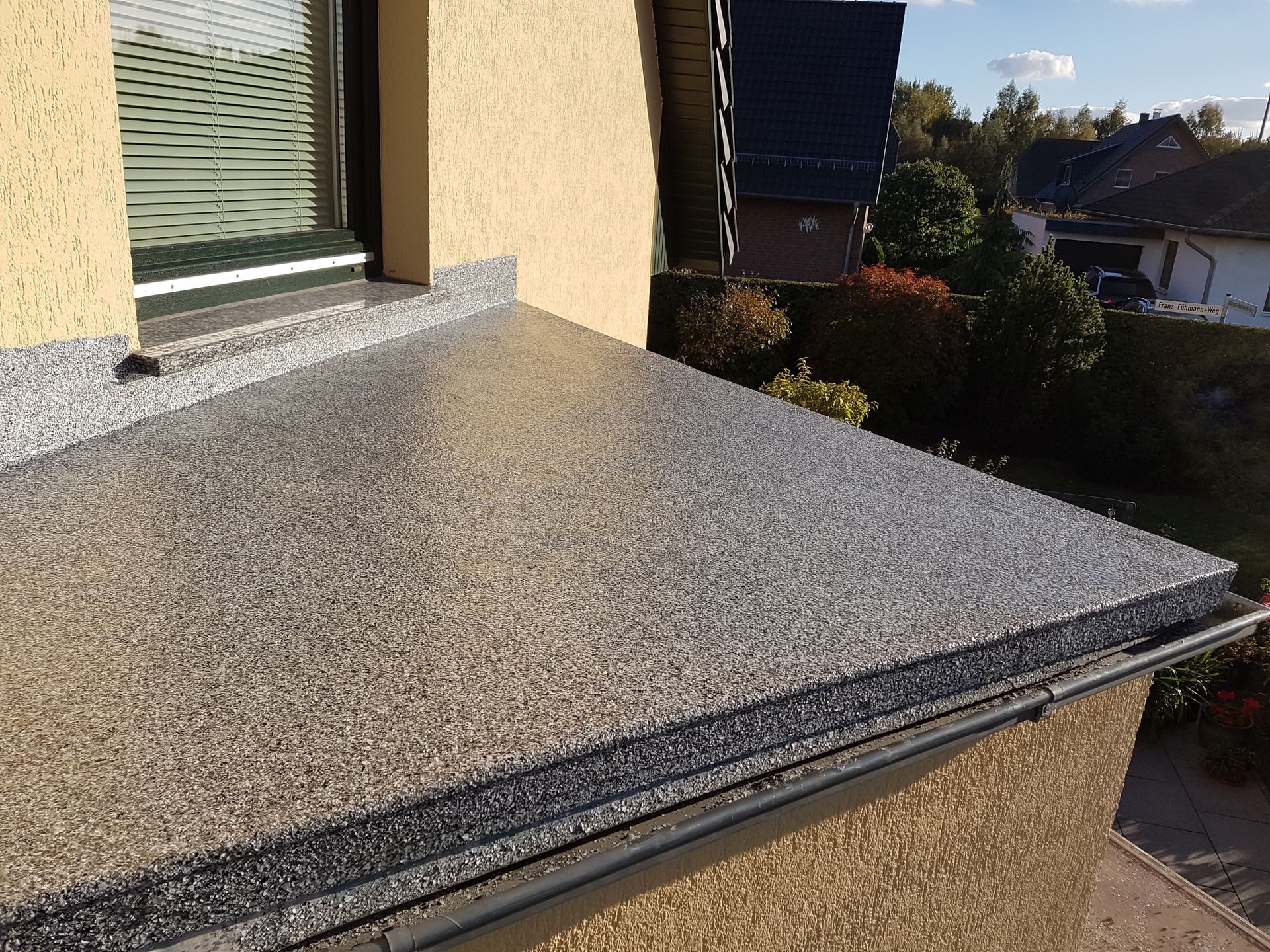 Nahaufnahme eines Balkons mit gesprenkelter grauer Oberfläche, neben einem Fenster und einer hellgelben Wand, mit einer Dachrinne am Rand.