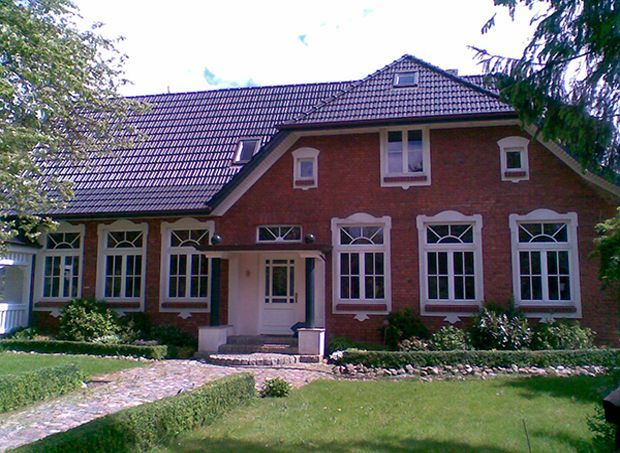 Ein Haus aus roten Backsteinen mit weißen Zierelementen und einem dunklen Ziegeldach, ein Gehweg und grüne Büsche.