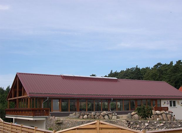 Ein Gebäude mit bordeauxrotem Metalldach und Holzfachwerk. Lange Fensterreihe, davor ein Steinhof.