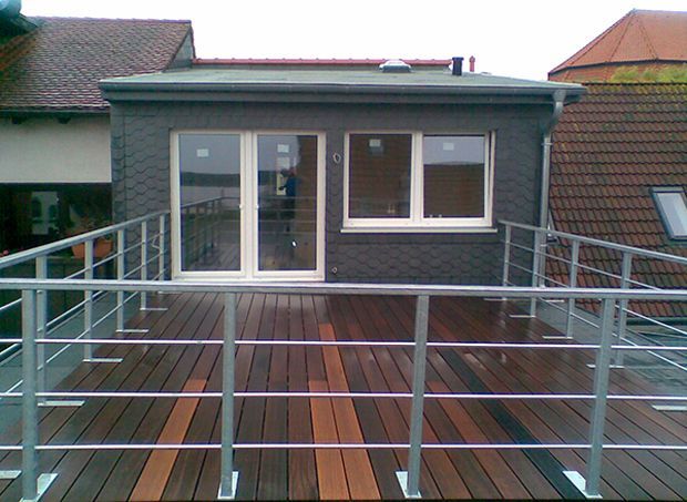 Dachterrasse mit Metallgeländer und Holzboden. Im Hintergrund ein dunkelgraues Gebäude mit Fenstern und einer Tür.