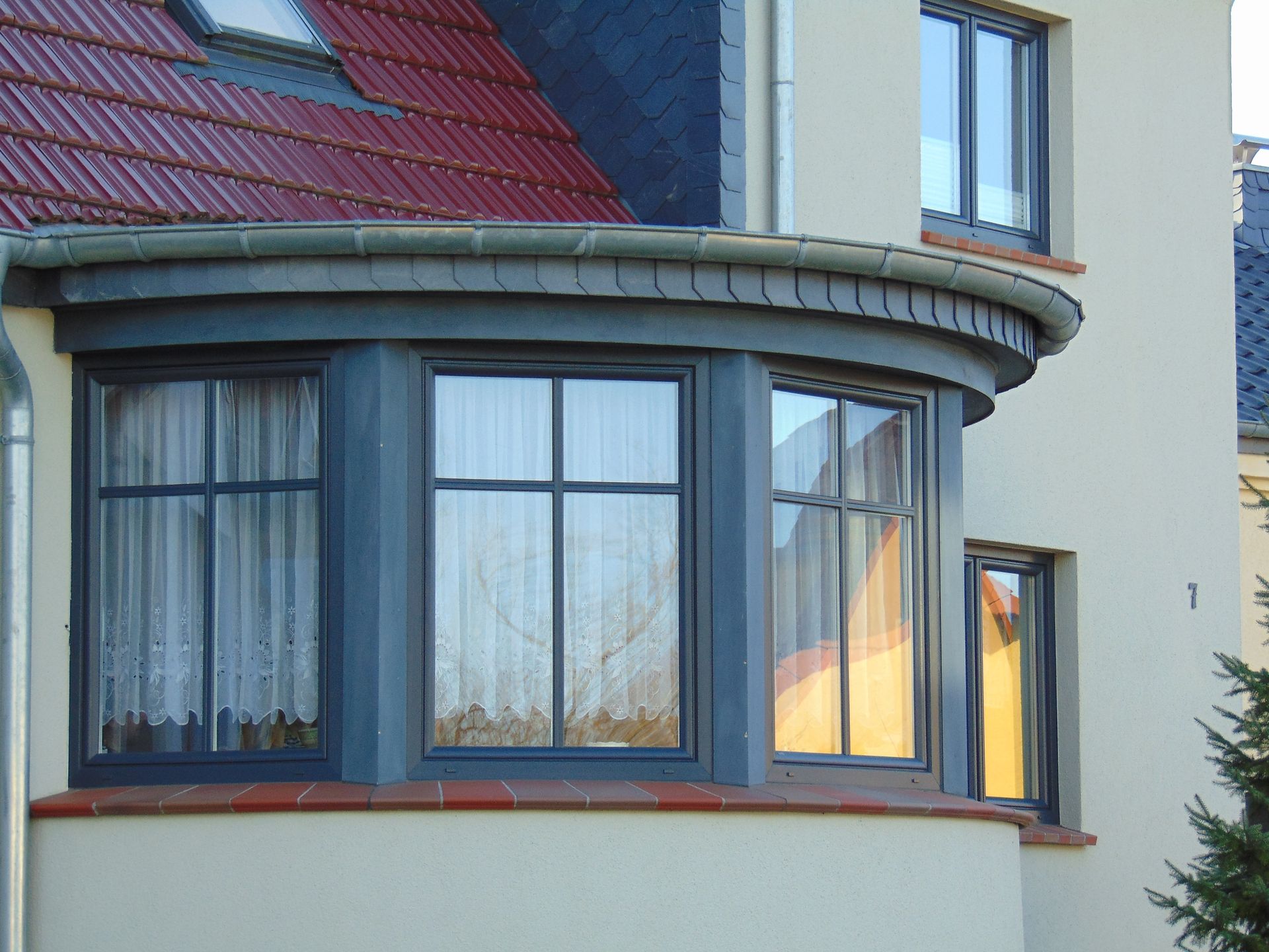 Gebogener Erker mit dunkelgrauen Rahmen an einem beigen Gebäude mit rotem Ziegeldach.