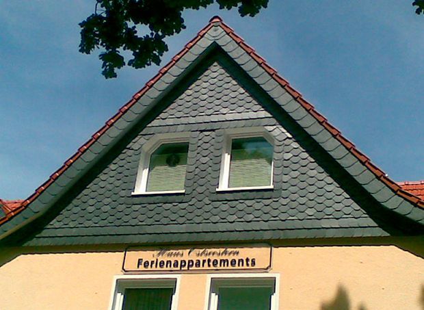 Dreieckiges Dach eines gelben Gebäudes mit grauem Schiefer und rotem Zierstreifen, zwei Fenstern und einem Schild mit der Aufschrift „Ferienappartements“.
