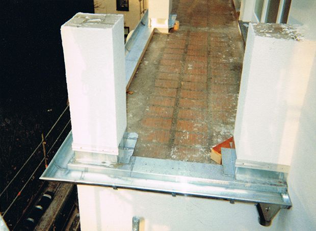 Ein schmaler Gehweg an der Außenseite eines Gebäudes, begrenzt von weißen quadratischen Säulen, Metallverzierungen und Ziegelboden.