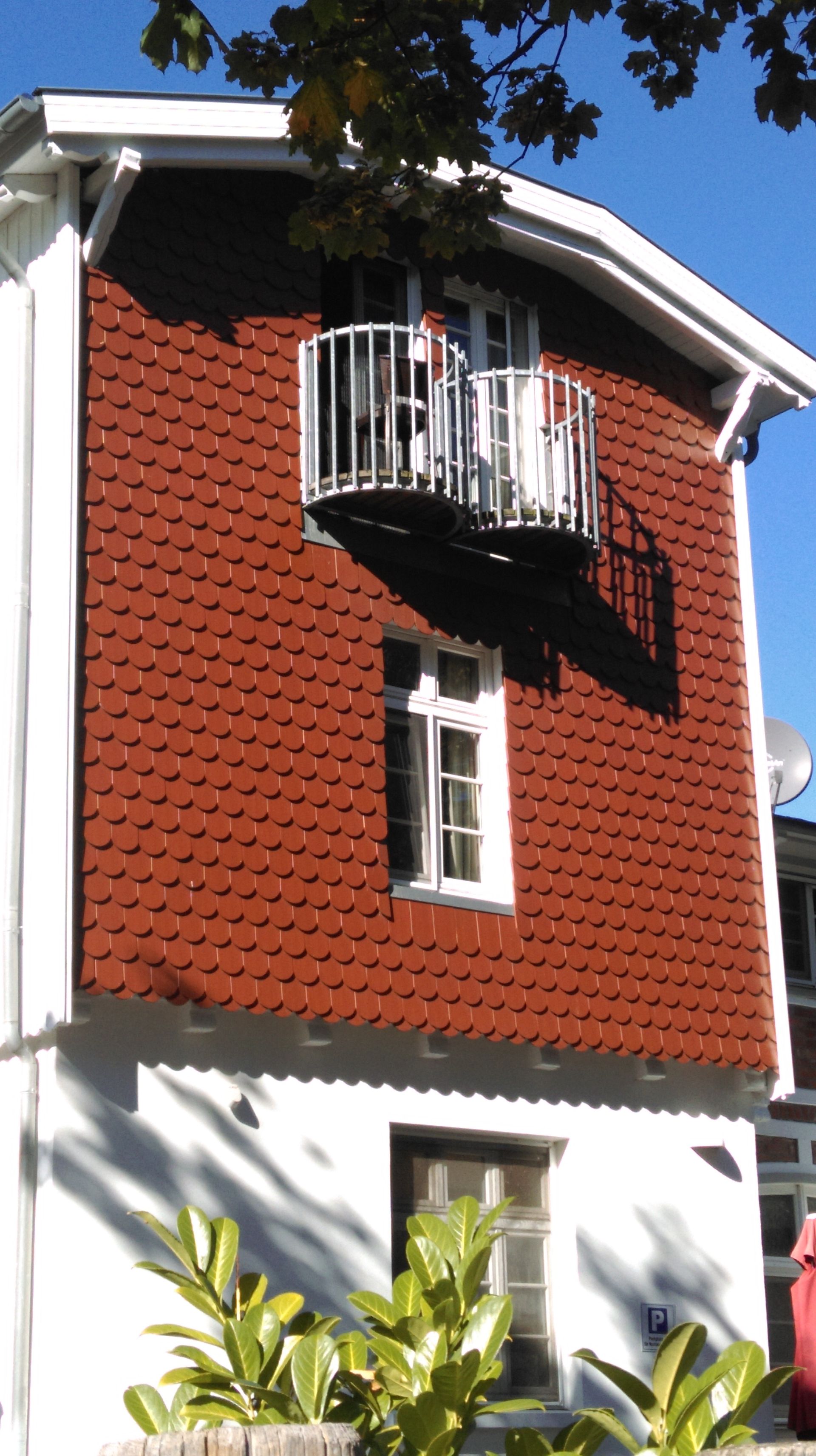 Ein rot gedecktes Gebäude mit weißen Zierelementen, kleinem Balkon und Fenstern. Grünes Laub im Vordergrund, blauer Himmel.