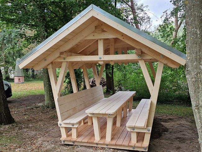 Hölzerner Picknickpavillon mit Sitzbänken und Tisch in einer Parkanlage.