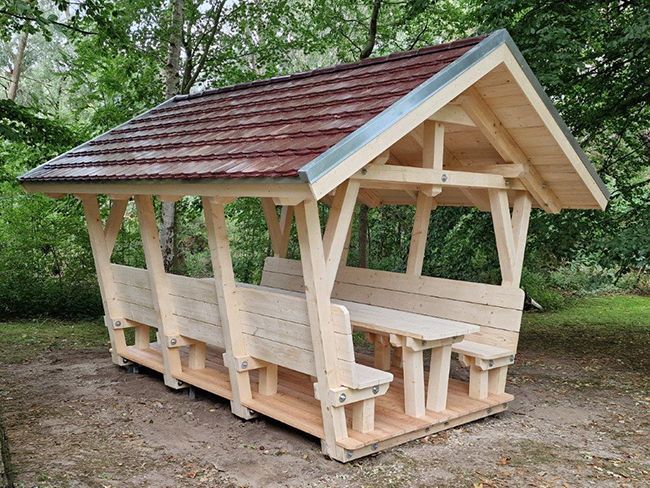 Hölzerner Picknickpavillon mit Sitzgelegenheiten und Tisch, braunes Ziegeldach, im Freien in einem Park aufgestellt.
