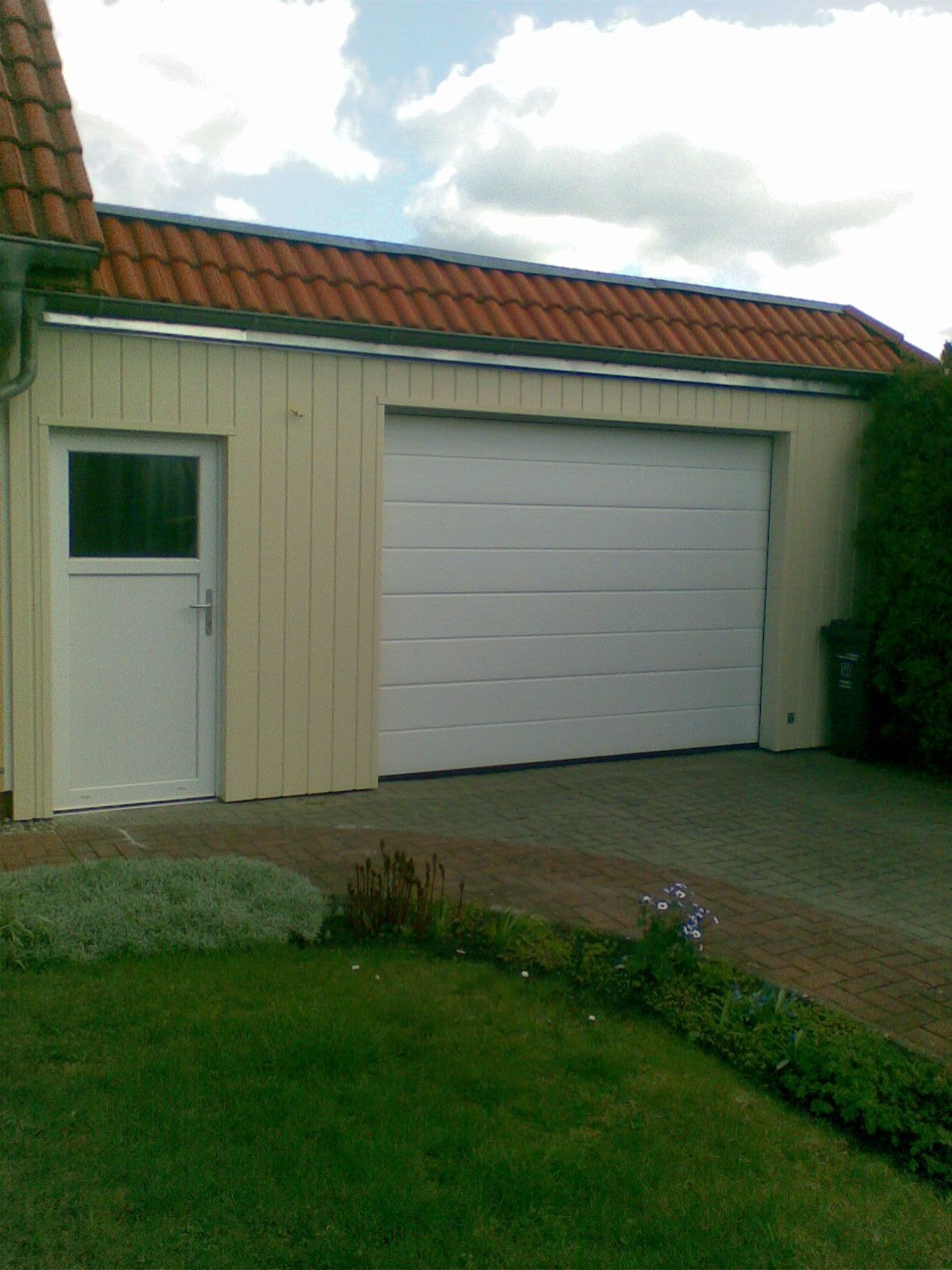 Beige Garage mit weißer Tür und Garagentor, rotem Ziegeldach und grünem Garten.