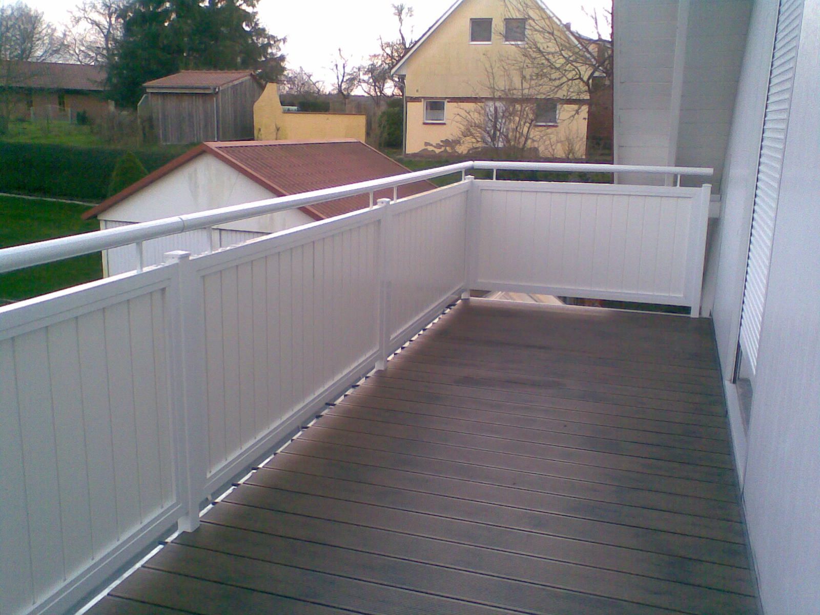 Balkon mit weißem Geländer und grauem Holzboden, mit Blick auf einen Hof und Häuser.