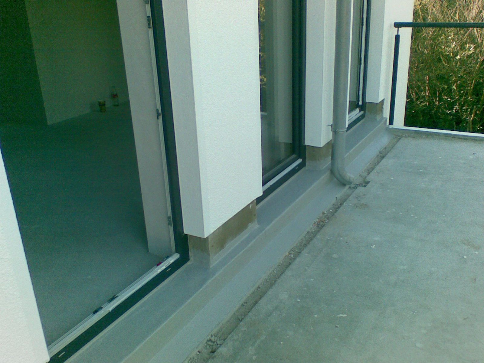 Balkon mit Glastüren, grauem Betonboden, weißen Wänden und einem Abflussrohr.