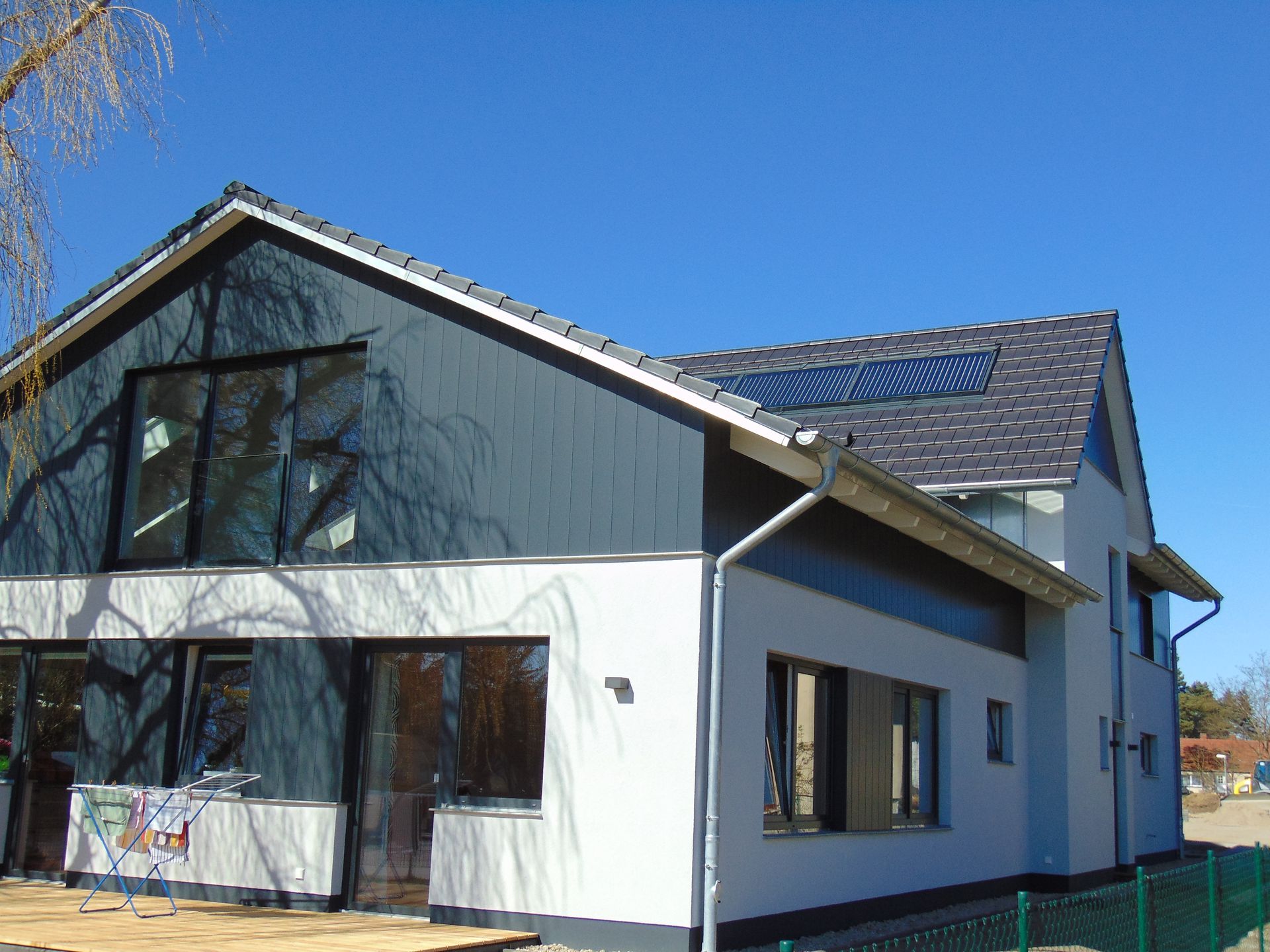 Modernes Haus mit grauem Dach und dunkelblauer Fassade, großen Fenstern und strahlend blauem Himmel.