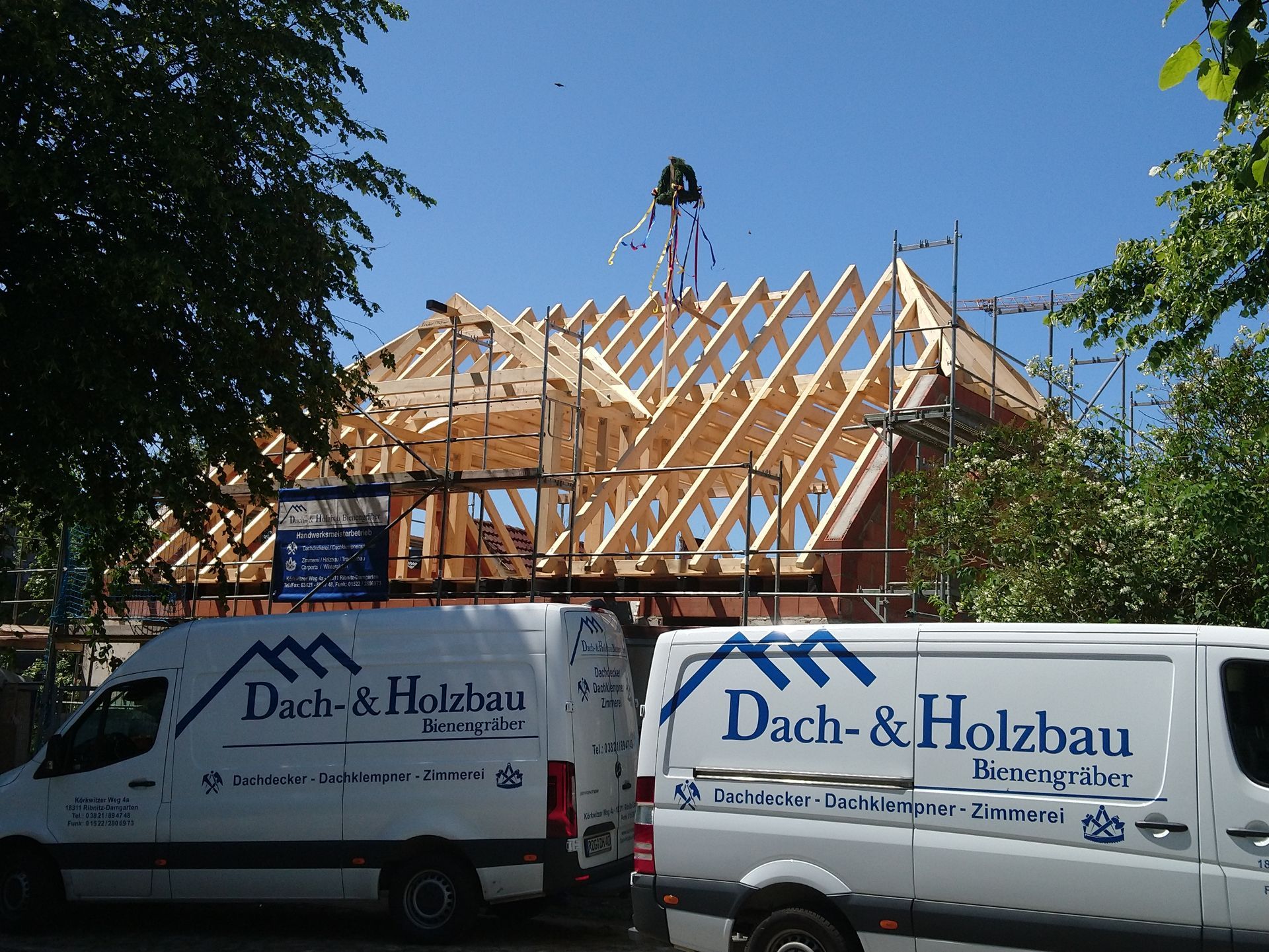 Zwei weiße Lieferwagen der Firma Dach & Holzbau vor einem im Bau befindlichen Haus; die Dachkonstruktion ist sichtbar.