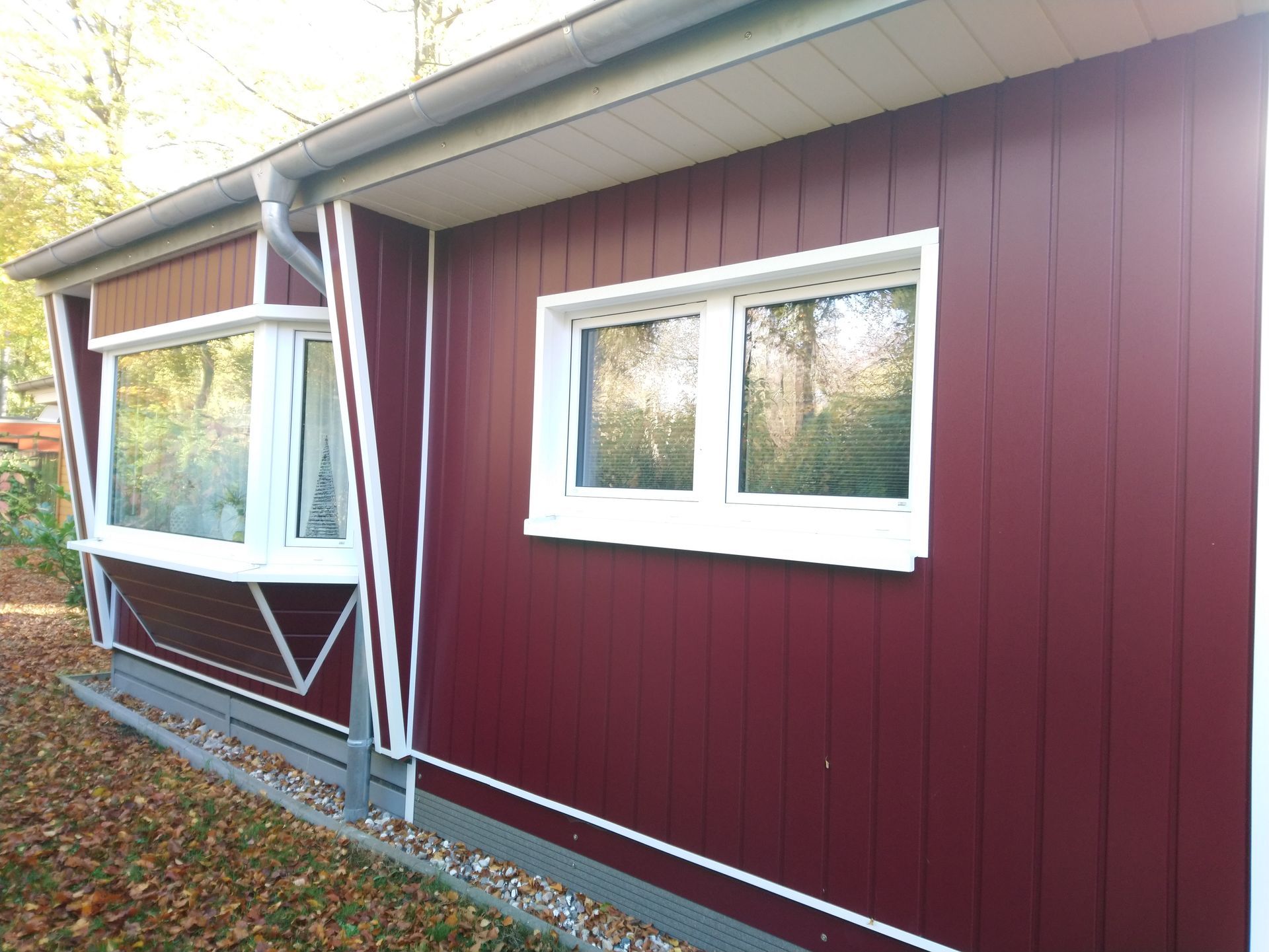 Ein rot verkleidetes Gebäude mit weißen Zierleisten um zwei Fenster. Herbstlaub auf dem Boden.