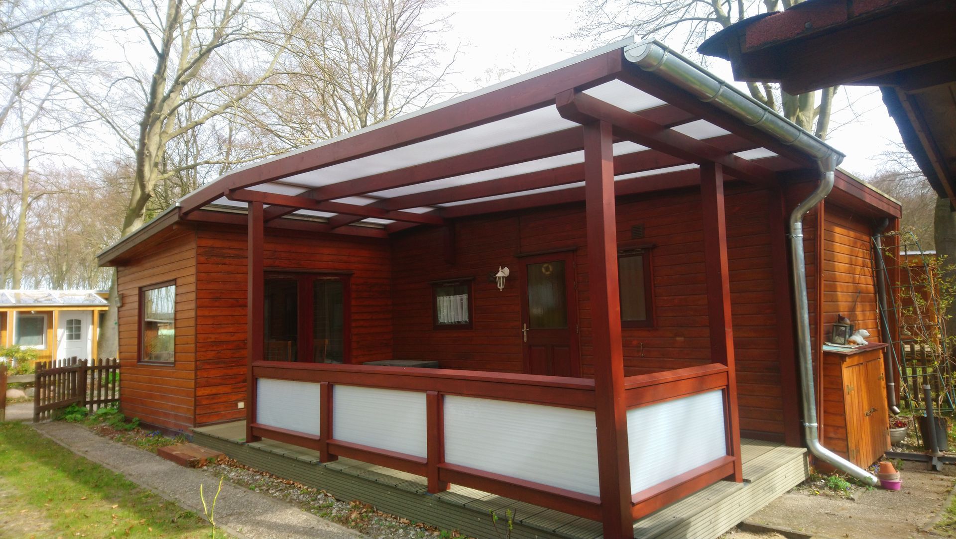 Holzhütte mit überdachter Veranda, rotbrauner Beize, weißem Geländer und transparentem Dach.
