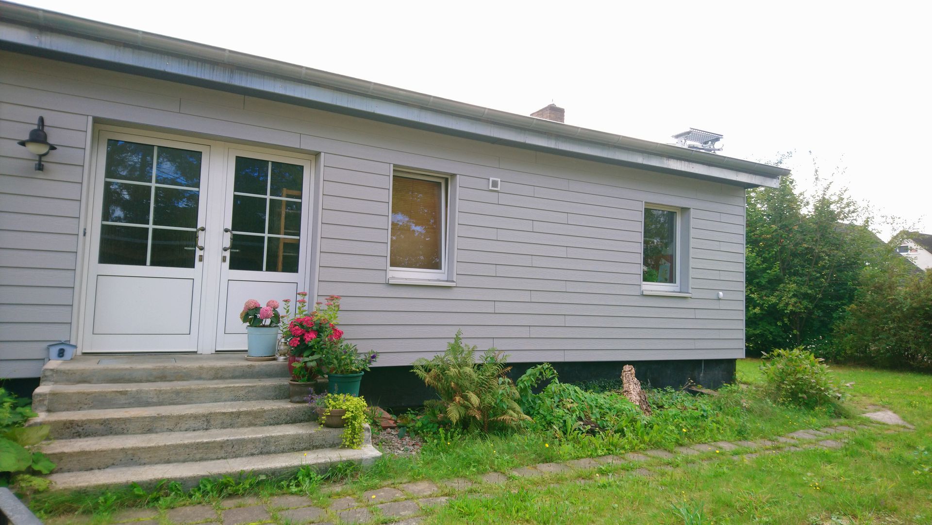 Graues Haus mit weißen Doppeltüren, Treppe und kleinen Fenstern. Pflanzen wachsen in der Nähe.