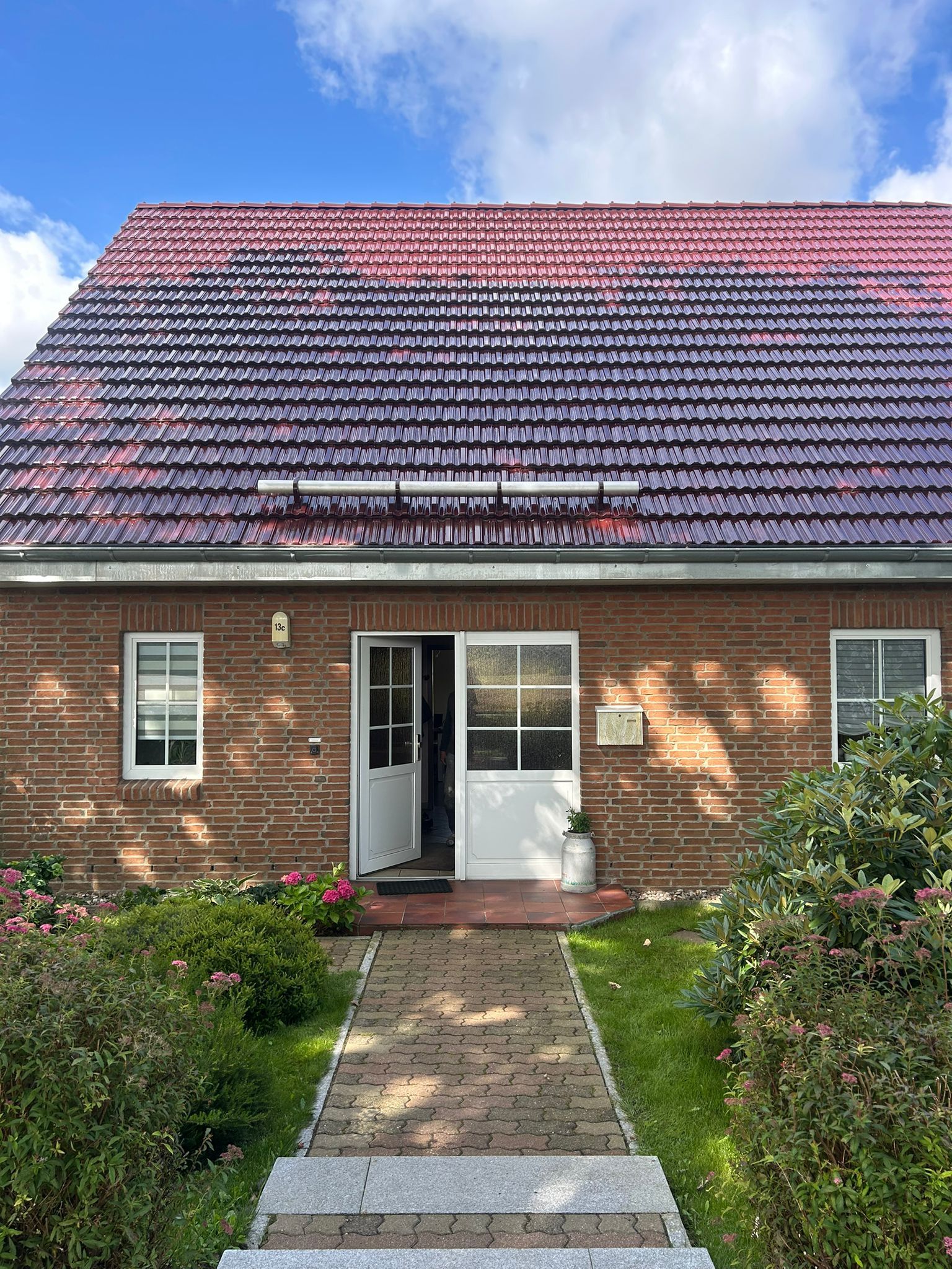 Backsteinhaus mit rotviolettem Dach, offener weißer Tür, Gehweg und Grünanlagen. Strahlend blauer Himmel.