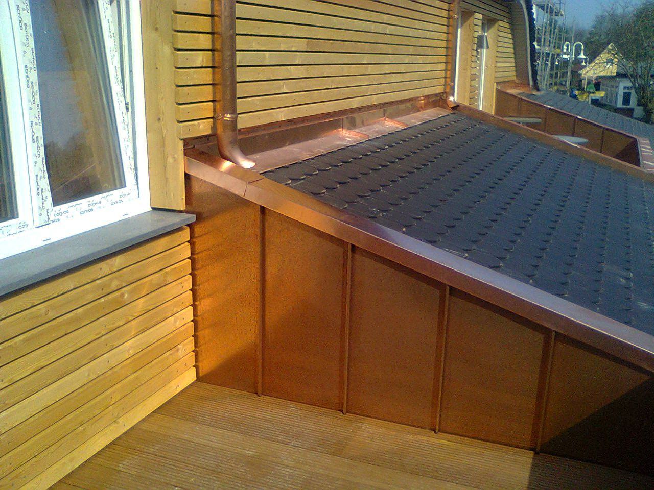 Kupferverkleideter Balkon mit brauner Holzverkleidung und schwarzem Dach. Regenrinne und Fenster sind sichtbar.