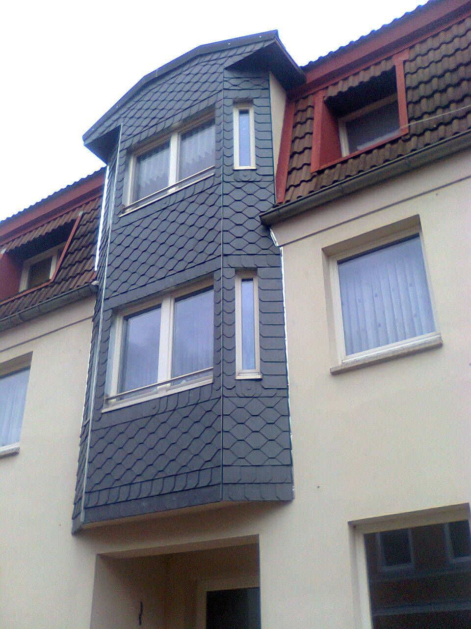 Die Fassade des Gebäudes ist mit dunkelgrauem Schiefer verkleidet und weist mehrere Fenster auf. Das rote Dach ist sichtbar.