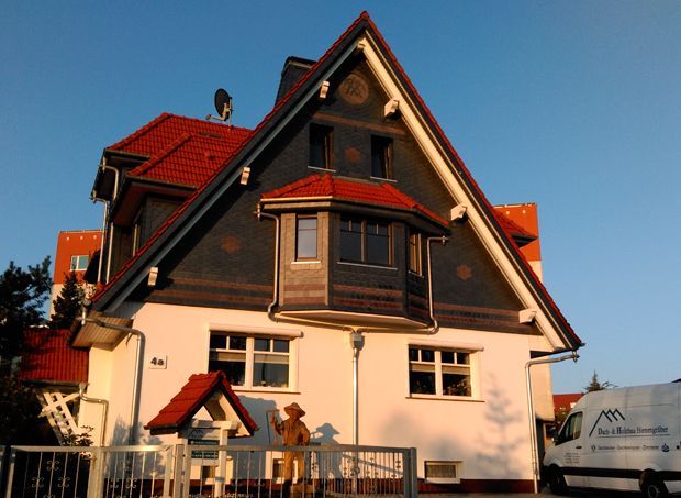 Haus mit rotem Dach und dunkler Fassade, Erkerfenster, weiße Wände, sonniger Tag.