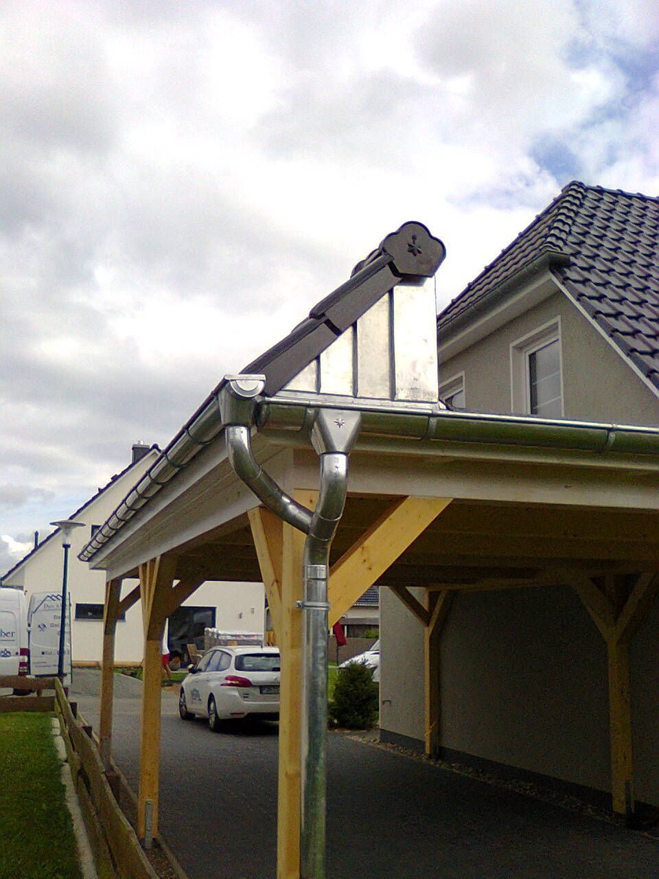 Carport mit grauen Dachziegeln, Holzrahmen und silberner Dachrinne, im Hintergrund bewölkter Himmel.