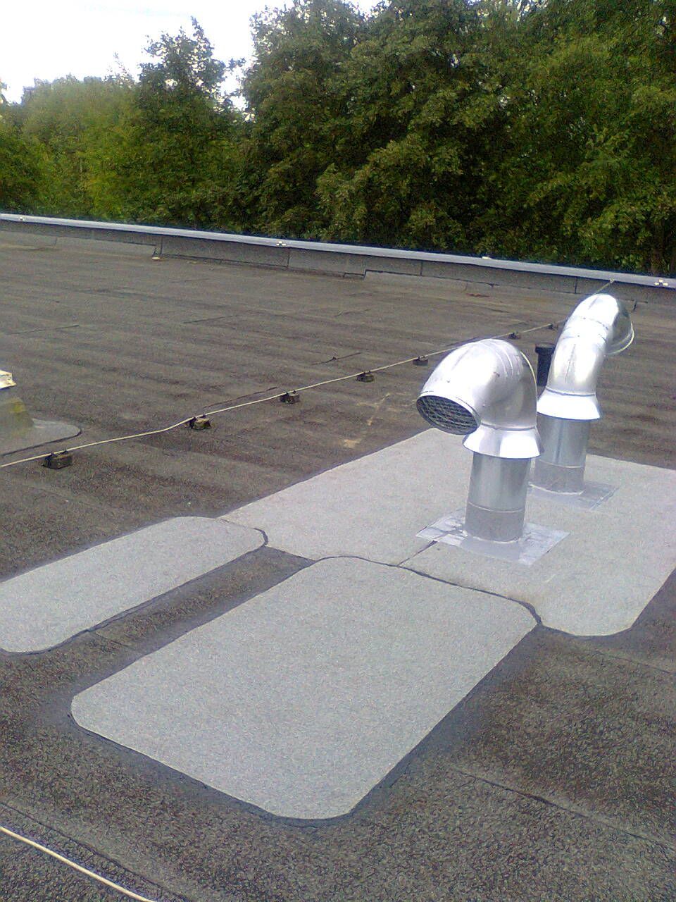 Schwarzes Dach mit zwei silbernen Auspuffrohren, grauen Flecken und Bäumen im Hintergrund.