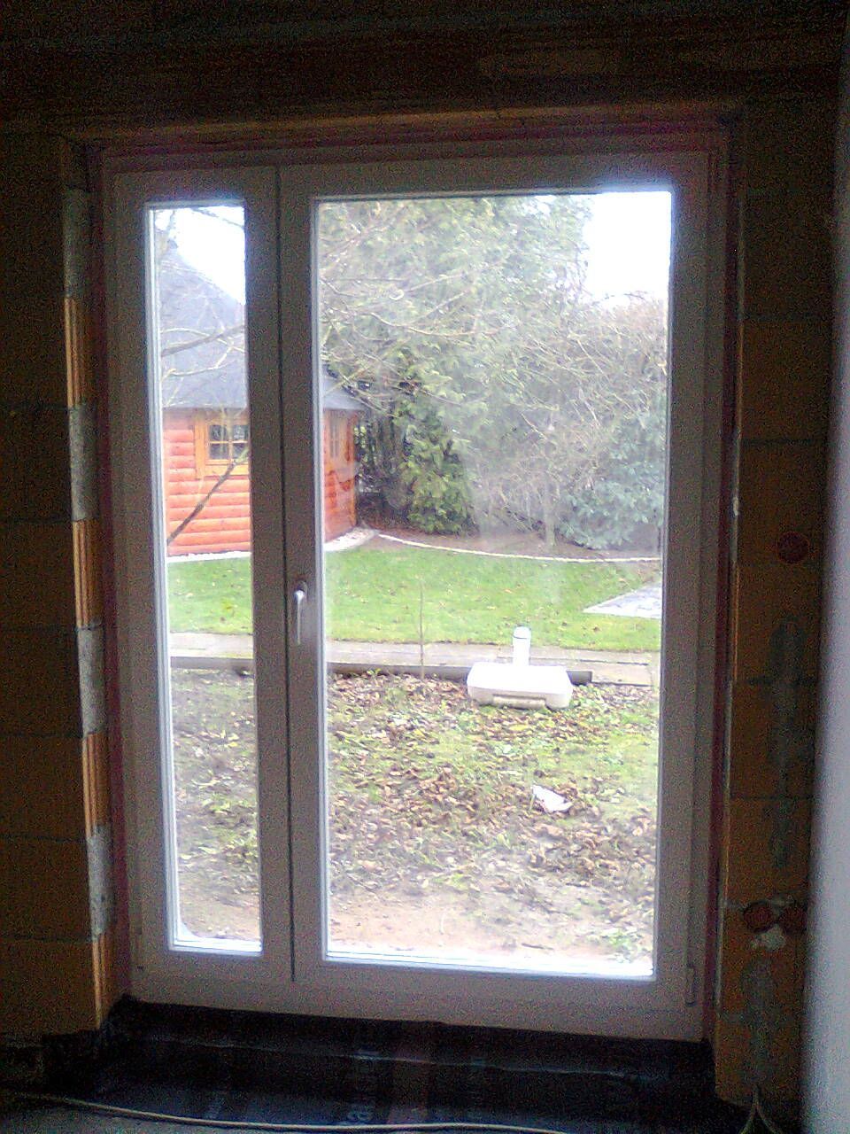 Fenster und Seitenwand mit weißem Rahmen; Außenansicht eines grasbewachsenen Hofes mit Bäumen und einem Gebäude.