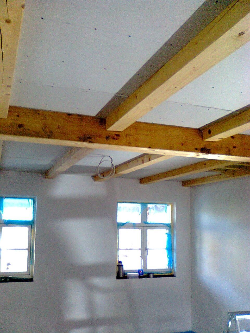 Innenansicht eines Zimmers mit sichtbaren Holzbalken, zwei Fenstern sowie weißen Wänden und Decke.