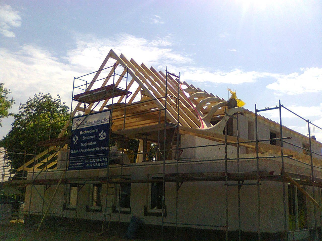 Ein im Bau befindliches Gebäude mit hölzerner Dachkonstruktion, umgeben von einem Gerüst, unter sonnigem Himmel.