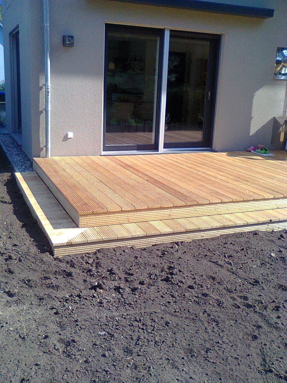 Eine Holzterrasse erstreckt sich von einem Haus mit Glasschiebetüren; im Vordergrund ist Schmutz zu sehen.