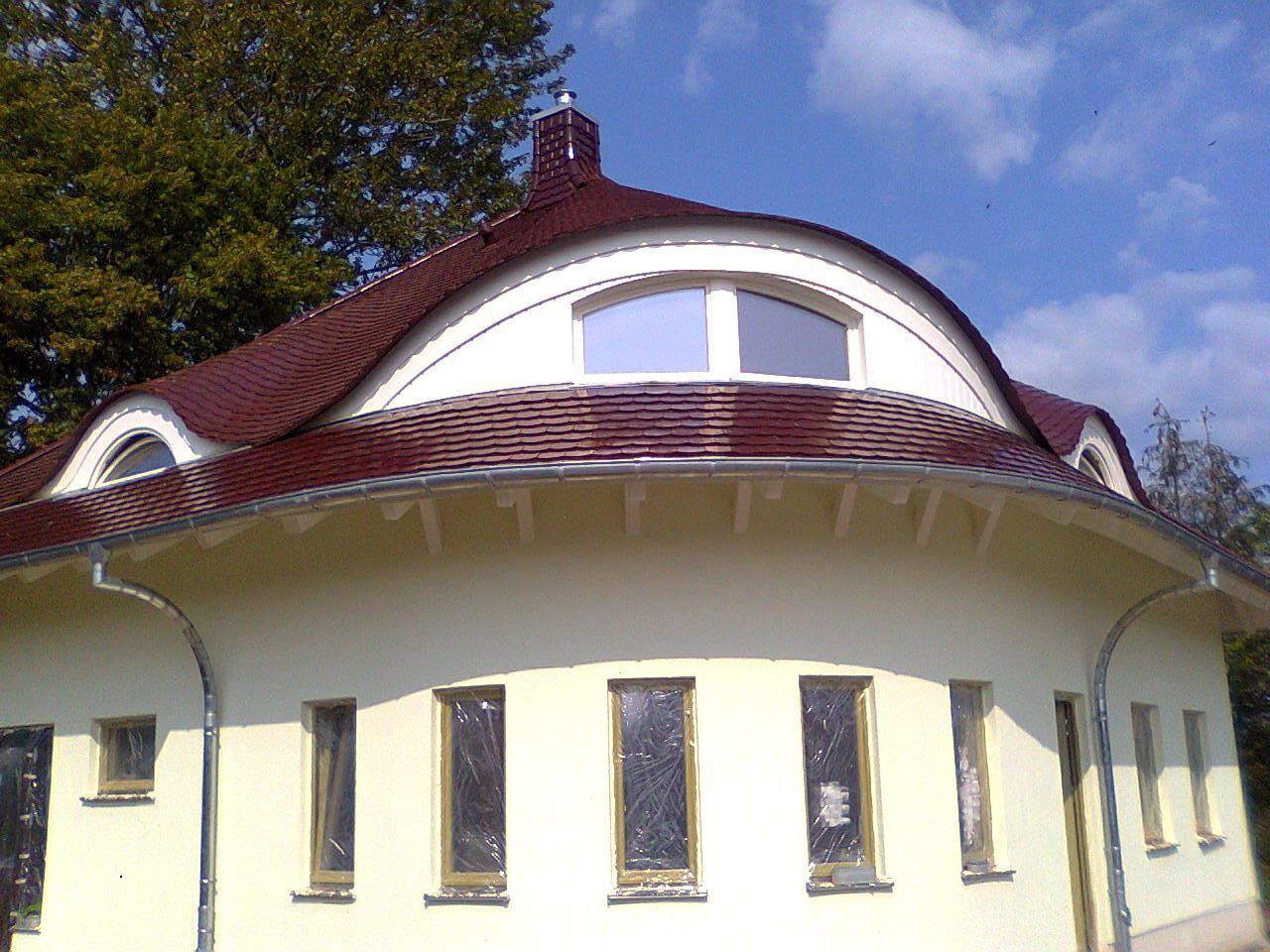 Rundes Gebäude mit bordeauxrotem Dach und cremefarbener Fassade. Mehrere Bogen- und rechteckige Fenster.