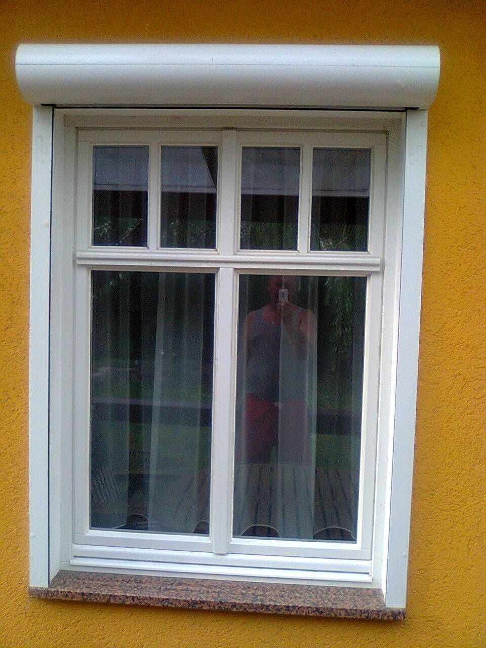 Weißes Fenster mit weißem Rollladen an einer gelben Wand, in dessen Glas sich eine Person spiegelt.
