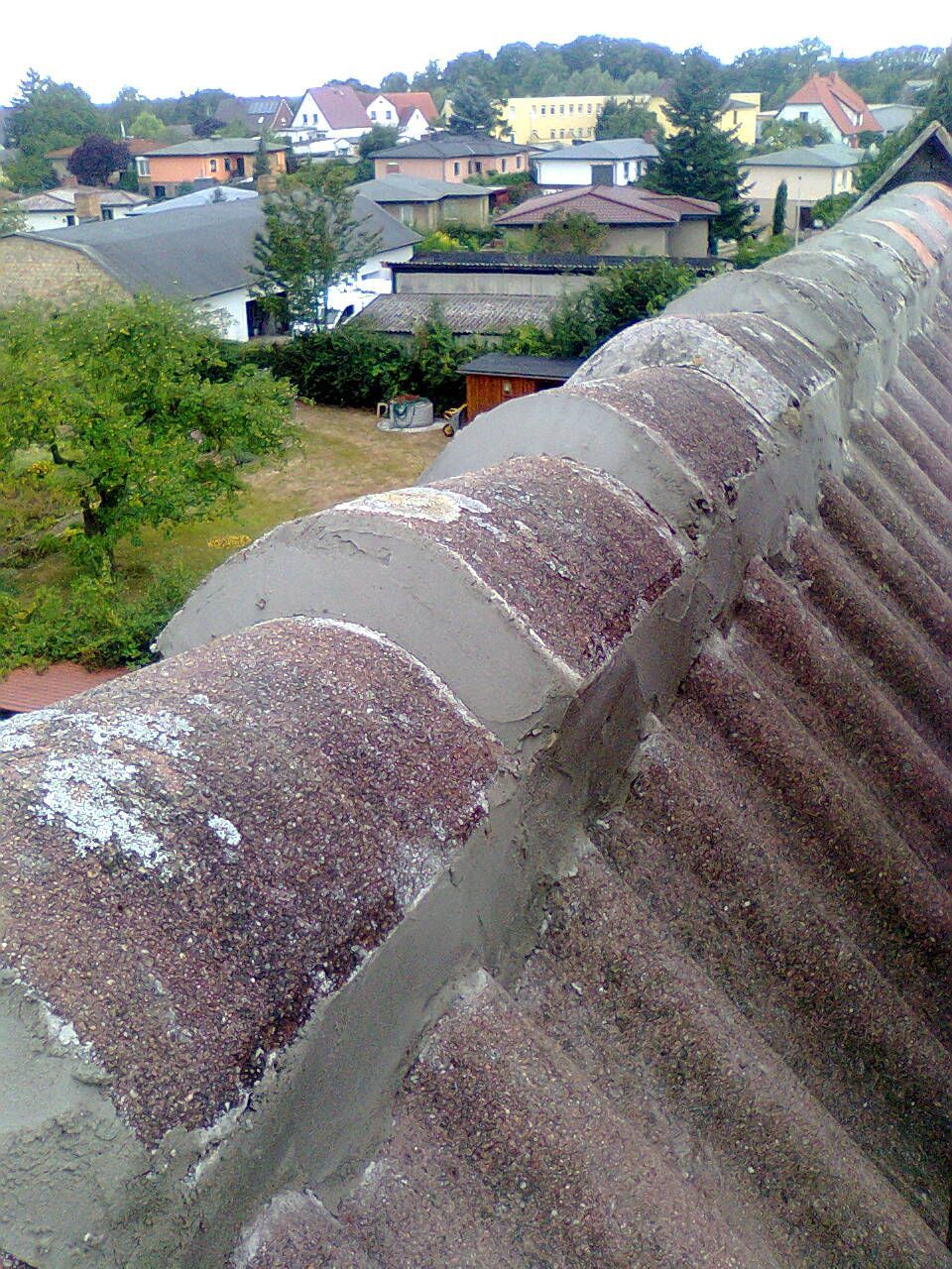 Nahaufnahme eines verwitterten, rotbraunen Wellblechdachs mit grauer Zementversiegelung, mit Blick auf ein Wohngebiet.