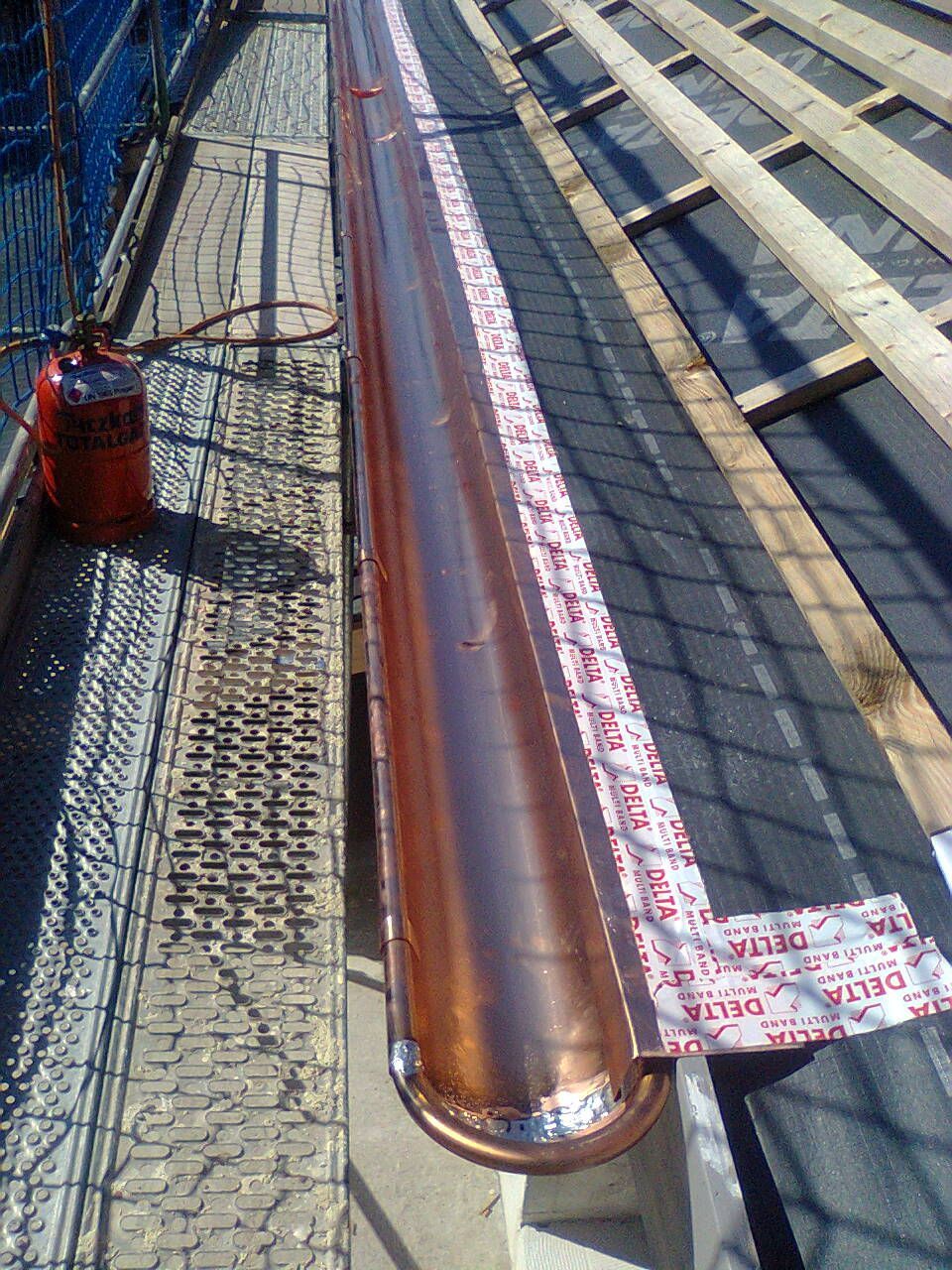 Eine Kupferrinne ist auf einem Dach mit sichtbarer Unterspannbahn und Gerüst montiert. In der Nähe befindet sich ein Propangastank.