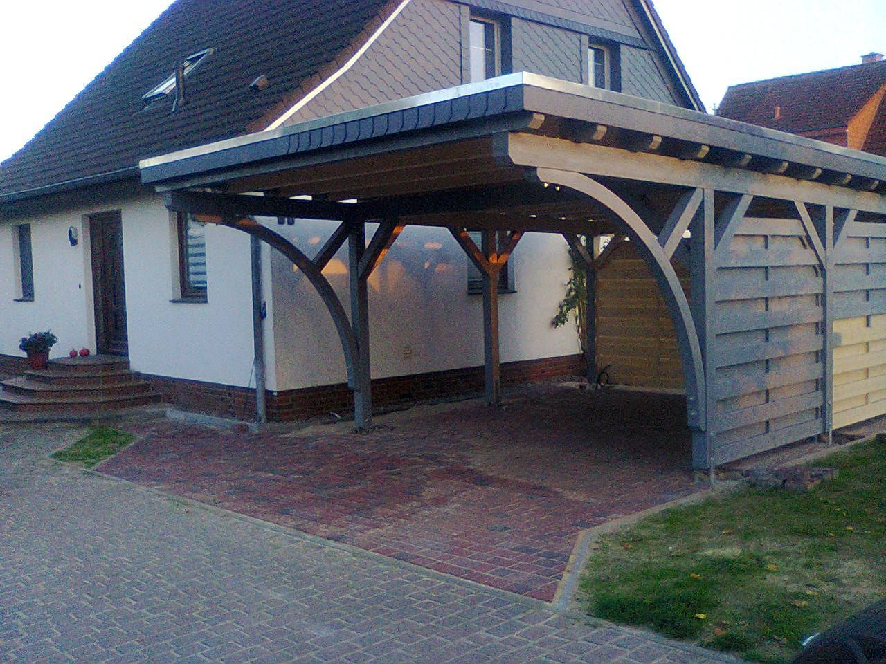 Ein Carport mit Holzstützen und gepflasterter Auffahrt ist an das Haus angebaut.