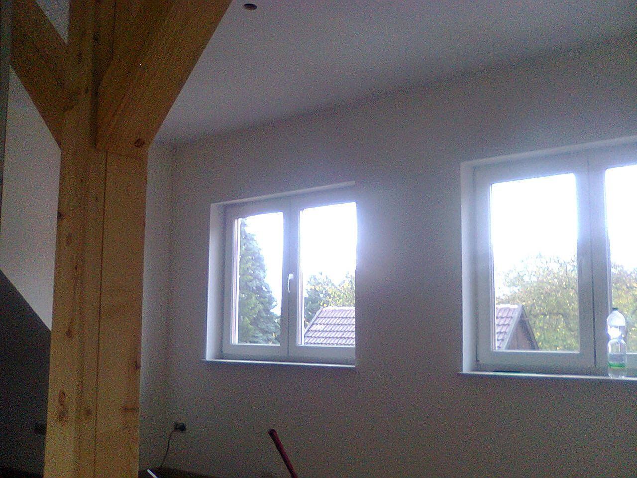 Innenansicht eines Zimmers mit zwei Fenstern, einem hölzernen Stützbalken und unverputzten Wänden.