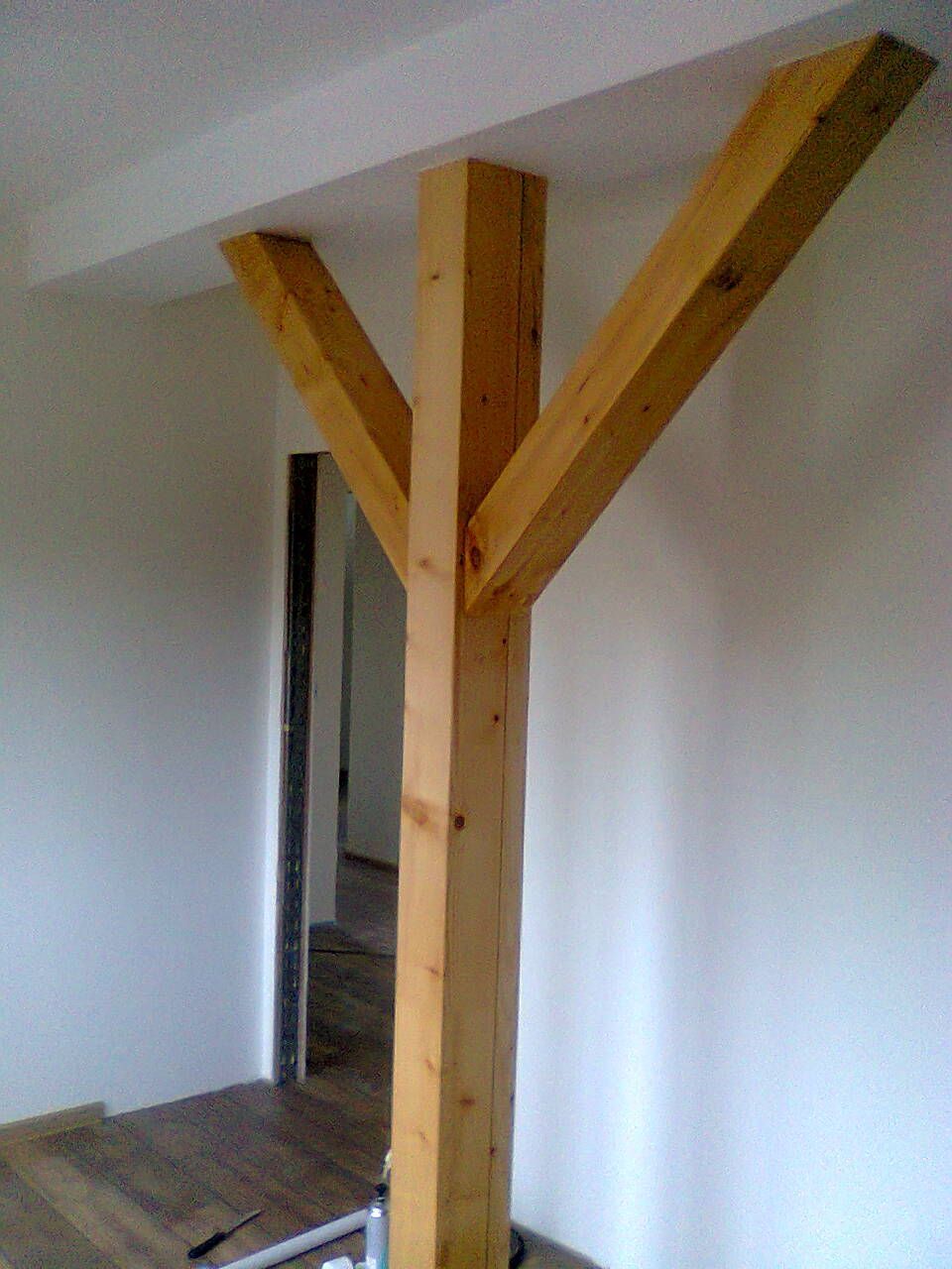 Eine hölzerne Stützsäule mit zwei schräg verlaufenden Balken, die mit der Decke verbunden sind, in einem Raum mit weißen Wänden und einer Tür.