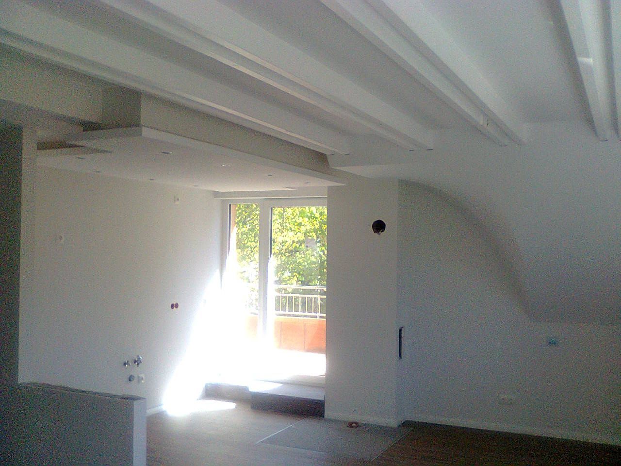 Innenraum mit weißen Wänden, schräger Decke und offener Tür zum Balkon mit Bäumen.